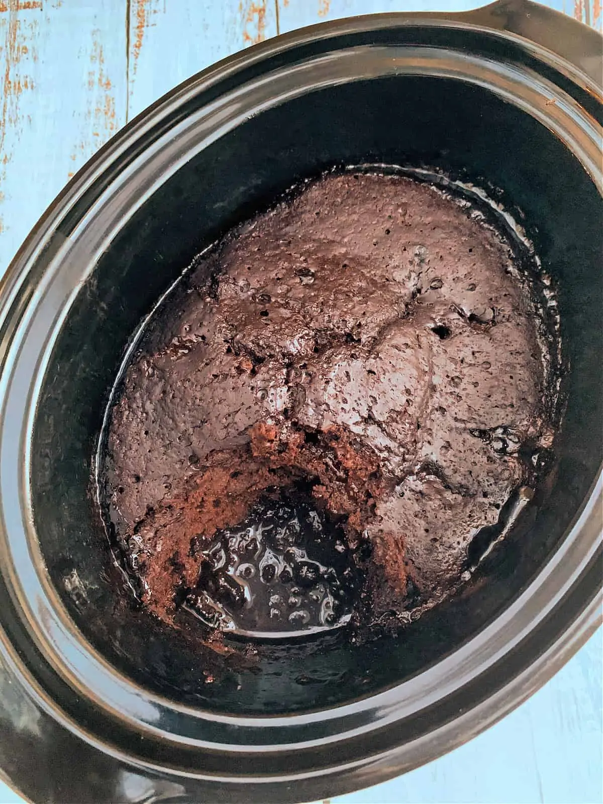 Chocolate sponge pudding with chocolate sauce in slow cooker pot, one portion removed to show the sauce.