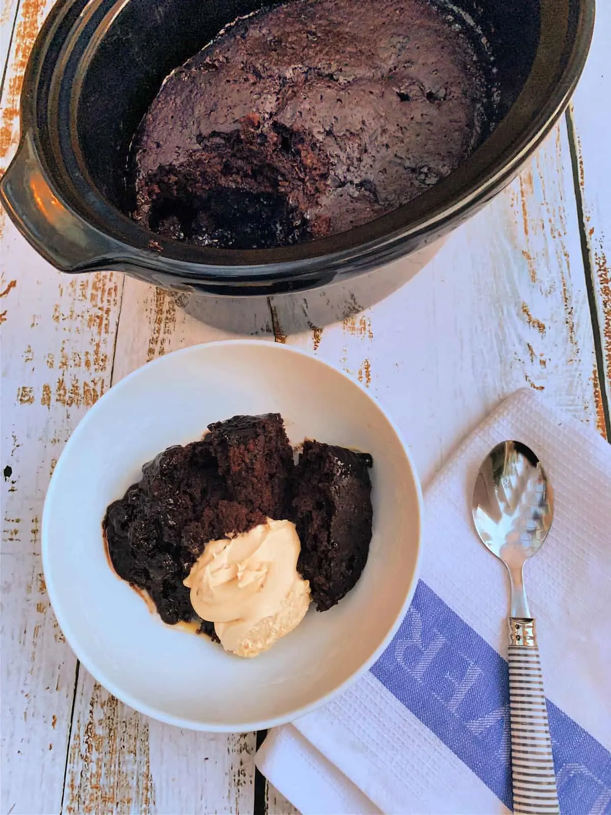 Slow cooker in background, bowl of chocolate lava cake with cream in front, with a spoon to the side.
