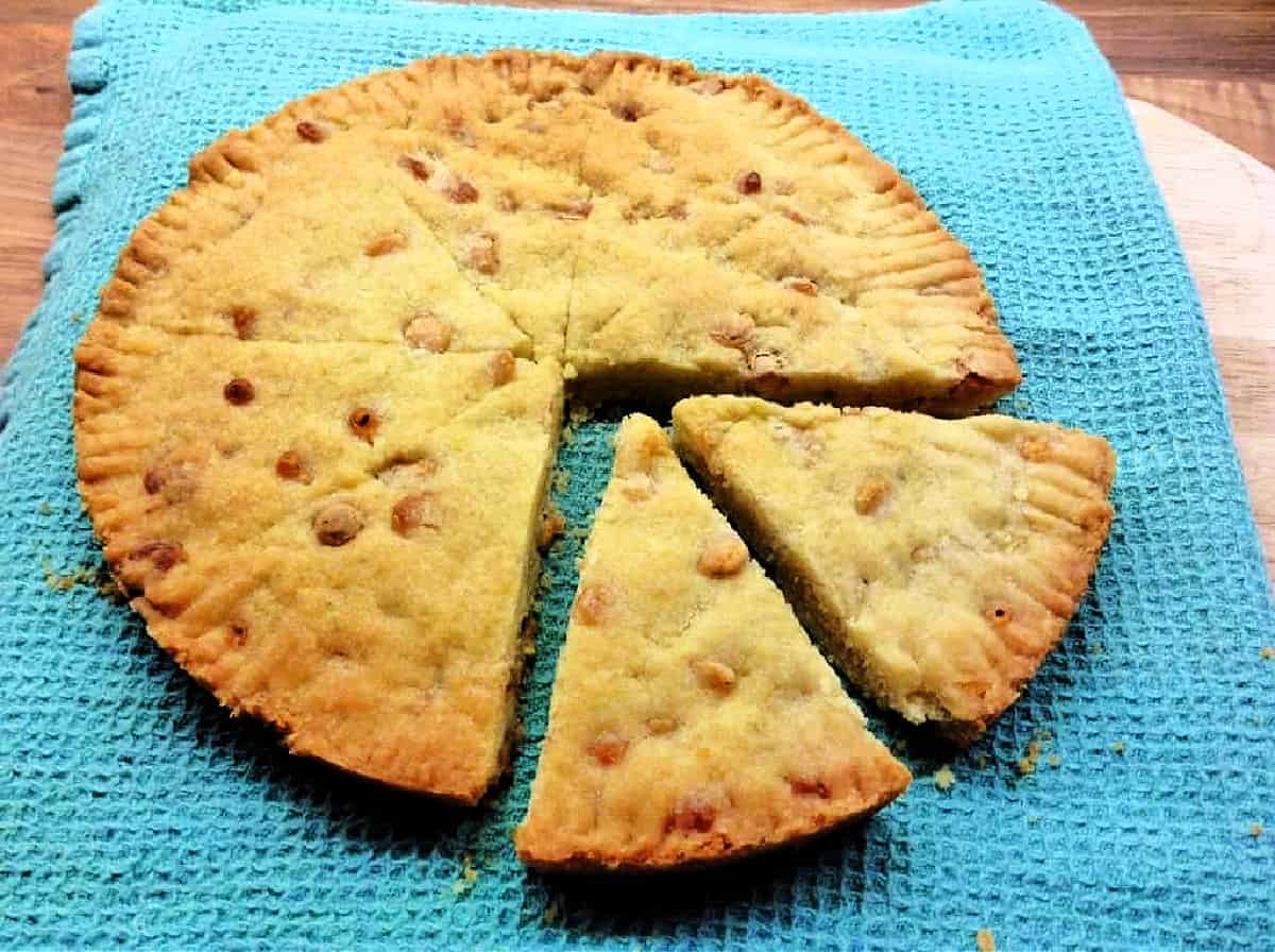 Round shortbread with white chocolate chips with two slices cut out, on light blue cloth.