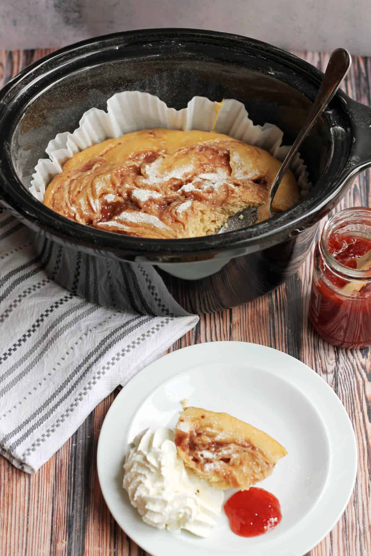 Cake in slow cooker pot, one portion on small plate with cream and jam.