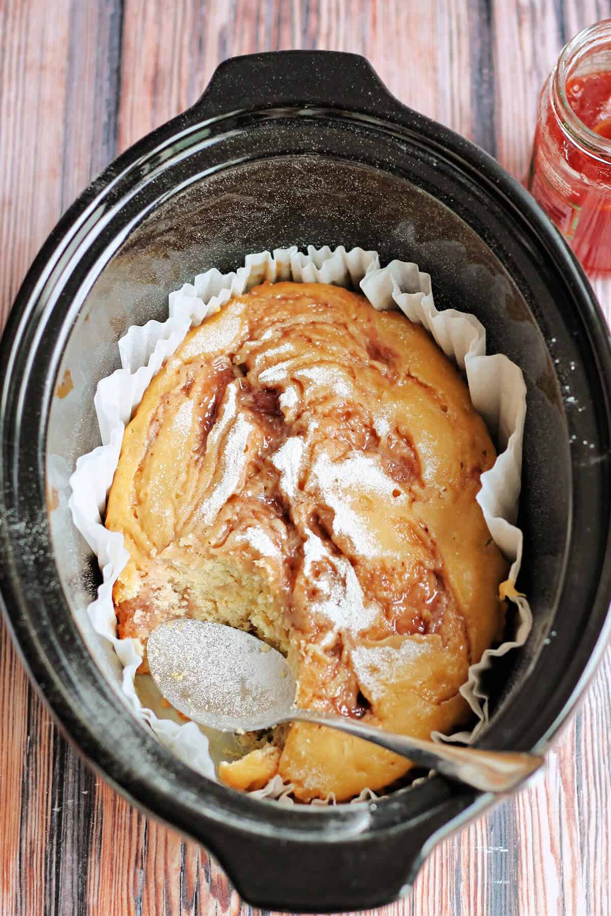A cake in a slow cooker pot dusted with icing sugar, with serving spoon.