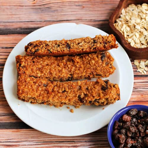 White plate with three oat and raisin cookie bars, with small bowls of raisins and oats on the side.