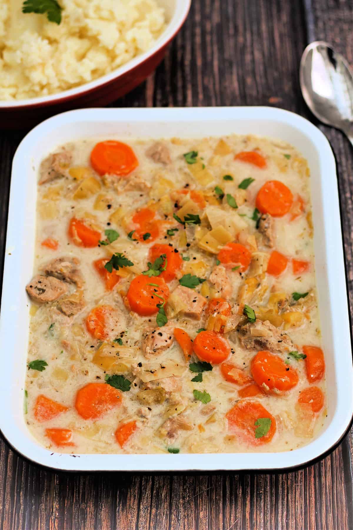 White serving dish with creamy pork casserole, with carrots and chopped fresh herbs.