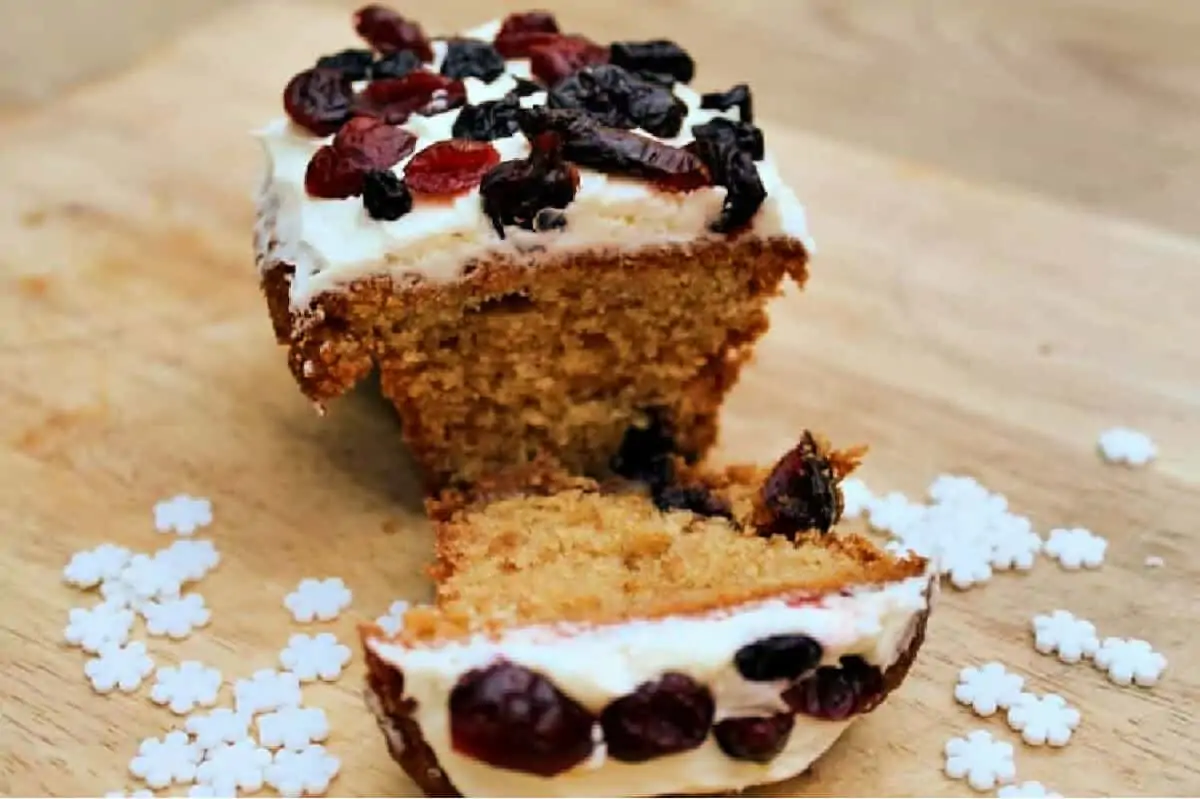 Small ginger loaf cake cut in half showing the soft inside, with white icing and berries on top, snow sprinkles around.