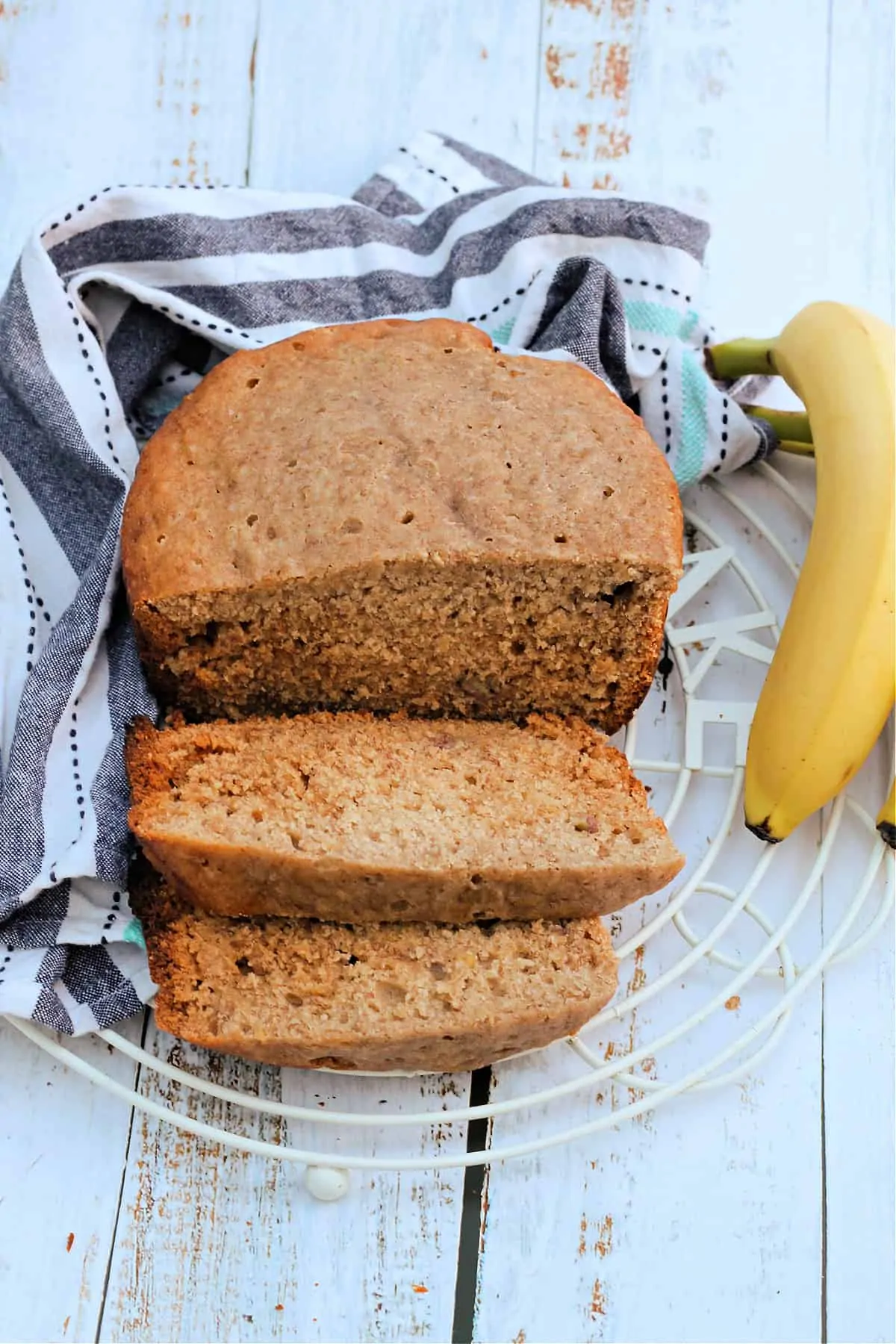 Banana bread on rack with two slices cut out, with bananas and a cloth behind.