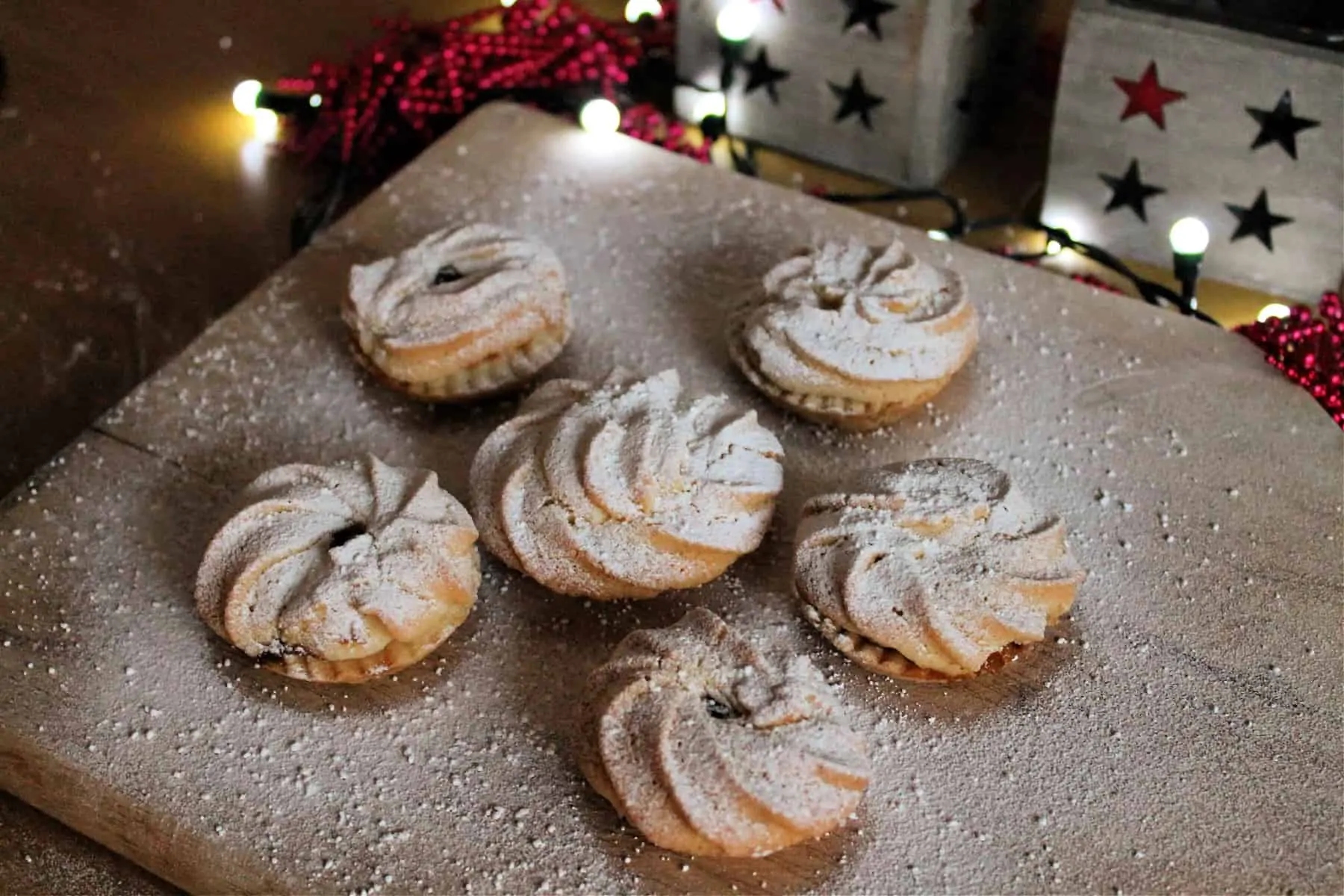 Close up of mince pies dusted with icing sugar on a wooden board, with fairy lights behind.