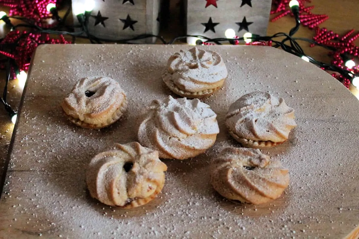 Close up of mince pies dusted with icing sugar on a wooden board, with fairy lights behind.