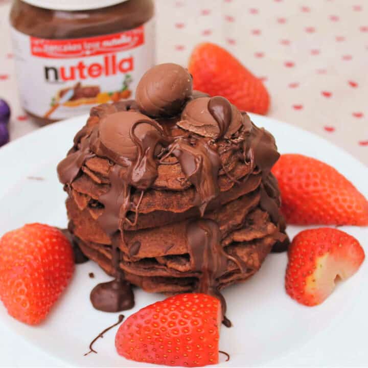 Stack of pancakes with chocolate drizzled over and strawberries around, jar of Nutella in background.
