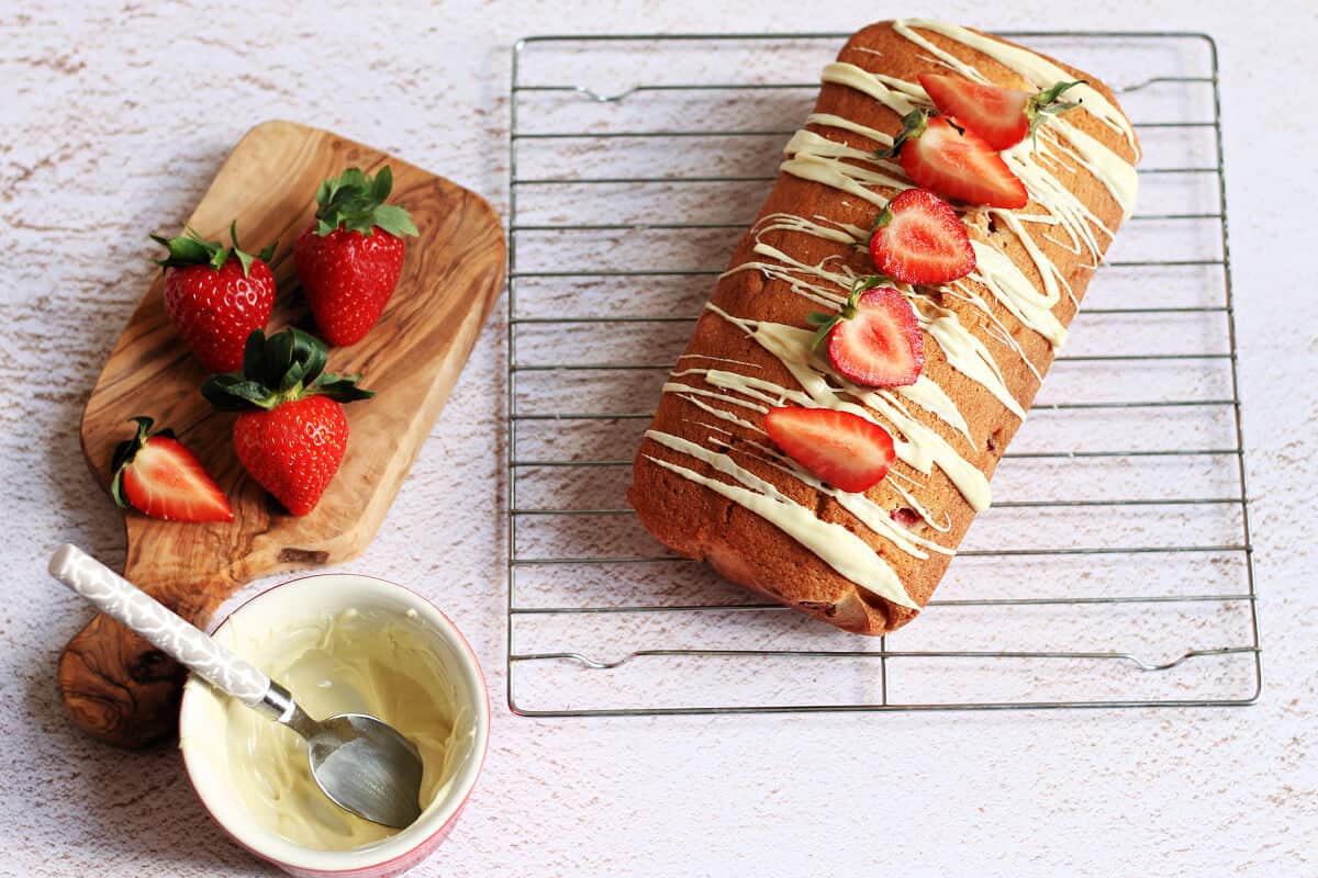 Loaf cake on a cooling rack, with white chocolate drizzled over it and chopped strawberries on top.