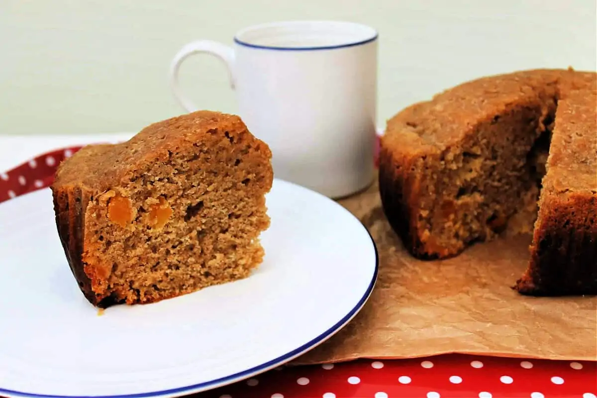 A slice of apricot cake on a white plate, white mug behind.