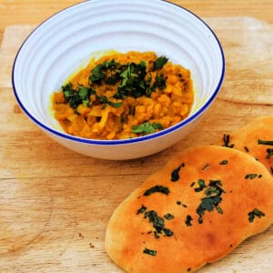 White bowl with yellow dal topped with chopped coriander on wooden board with coriander naan next to it.