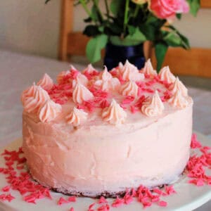 Strawberry cake with piped icing decoration on a table, roses behind in vase.