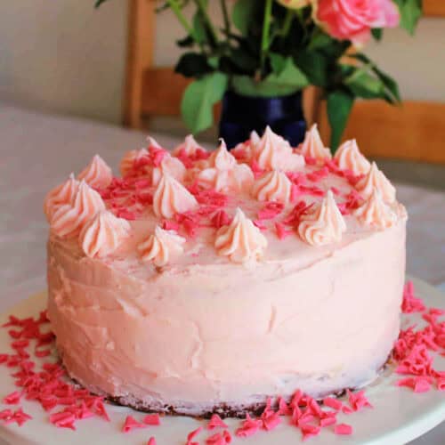 Strawberry cake with piped icing decoration on a table, roses behind in vase.