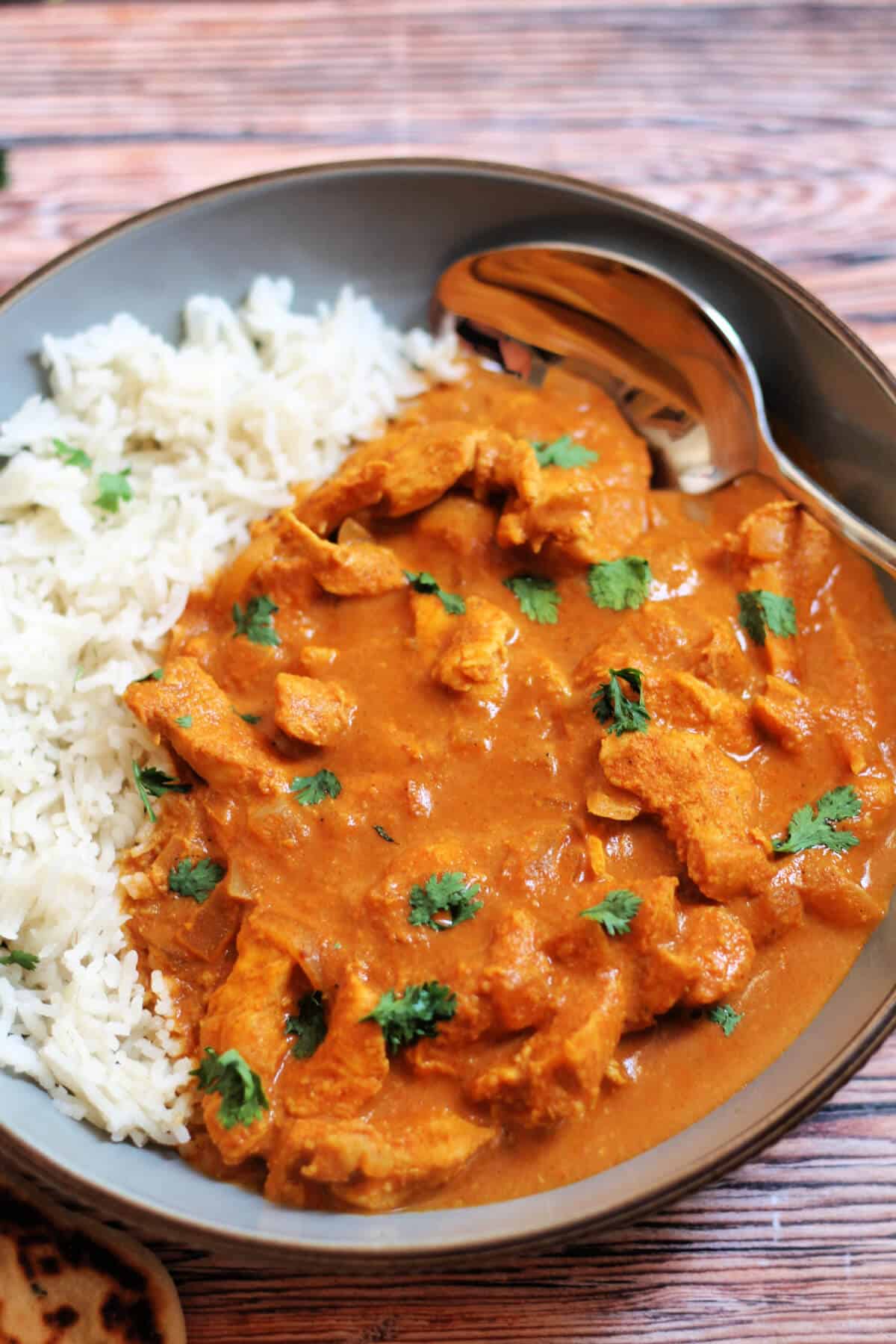 Close up of chunks of chicken in rich curry sauce, in bowl with rice and a spoon.