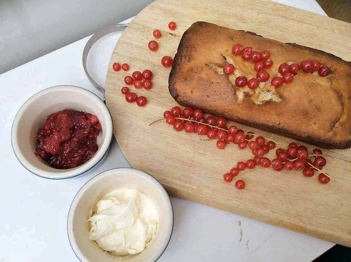 Loaf cake on a board, topped with redcurrants. Compote and yoghurt in pots to the side.