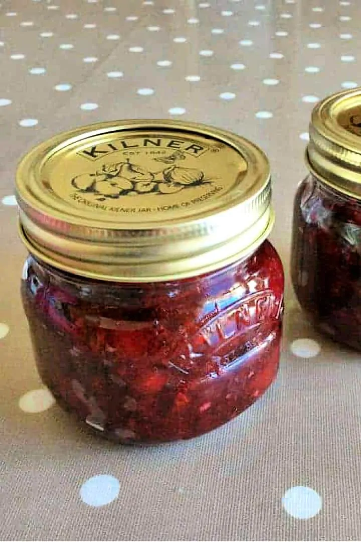 Close up of a small jar of red jam on a table with taupe table cover.