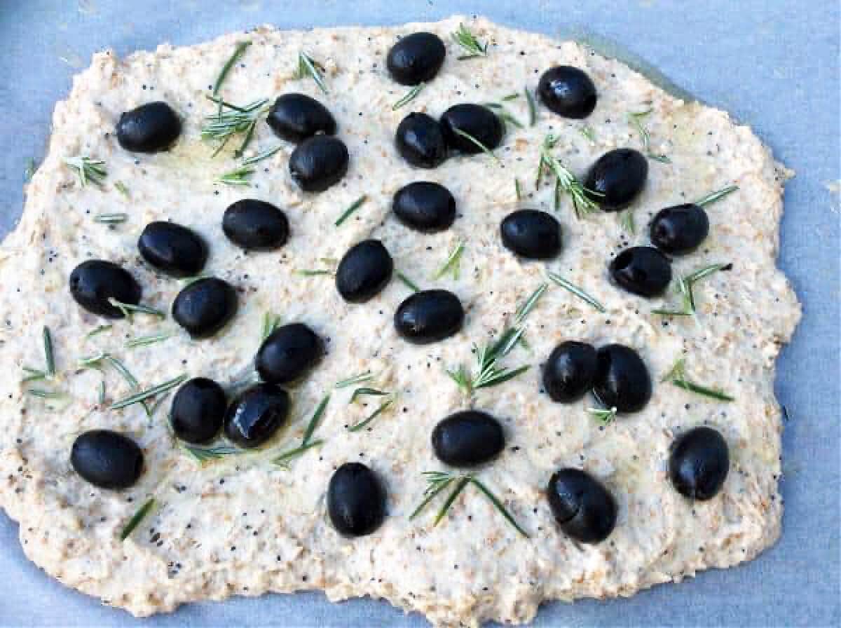 Focaccia bread dough on work surface, with black olives and rosemary.