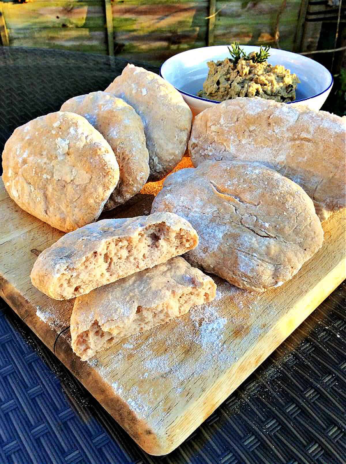 Pitta breads on a chopping board, one broken in half, with bowl of hummus at back.