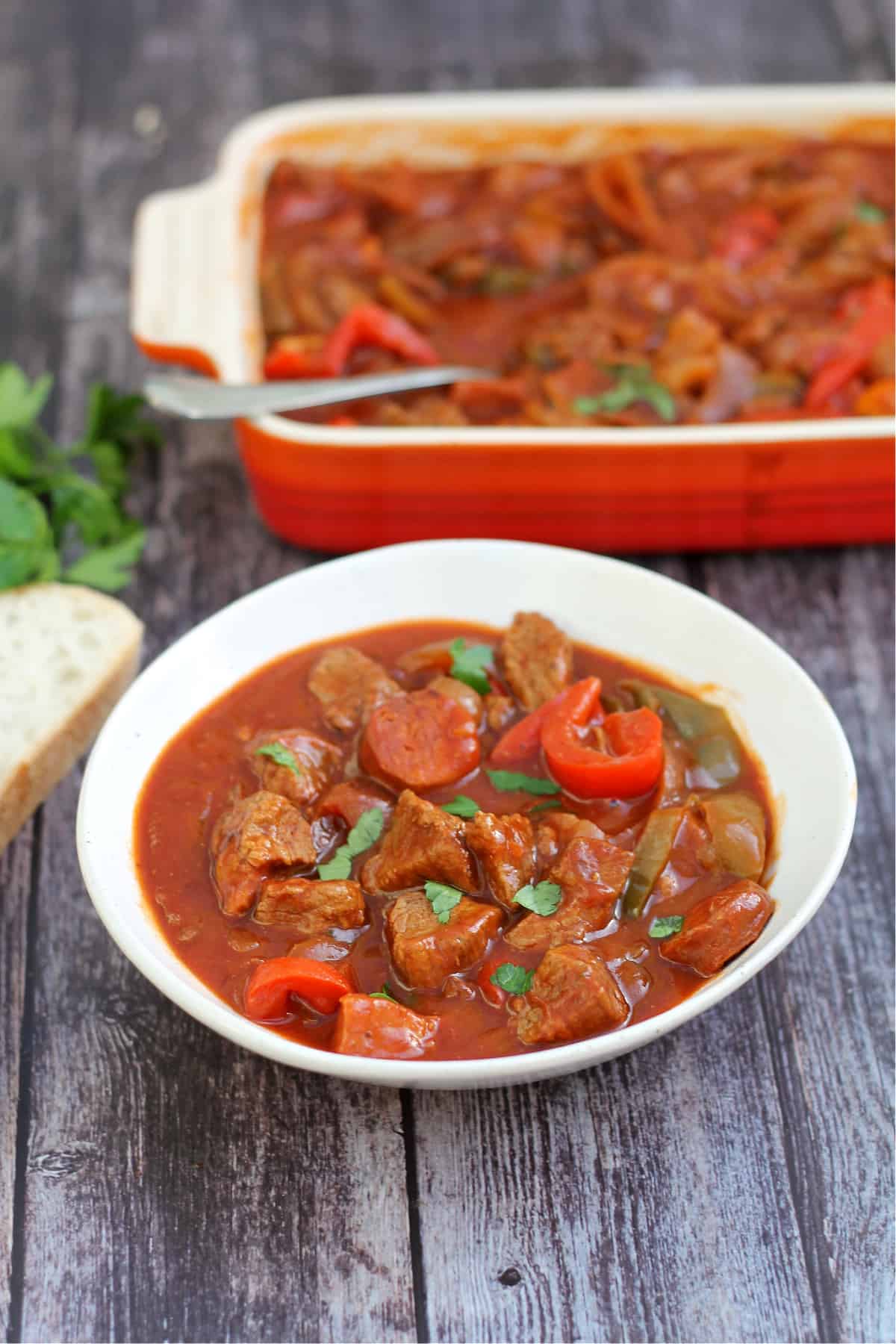 Bowl of beef stew with serving dish behind and bread to the side.