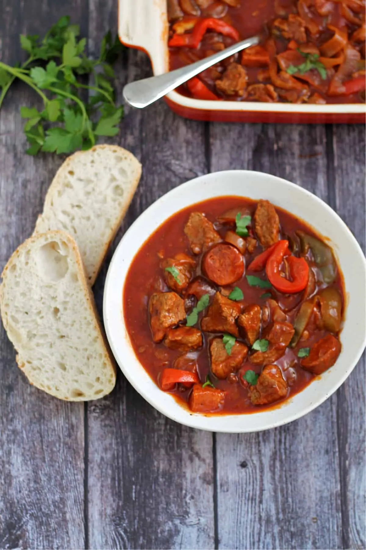 Bowl of beef casserole served with crusty bread on the side, with herbs and serving dish behind.