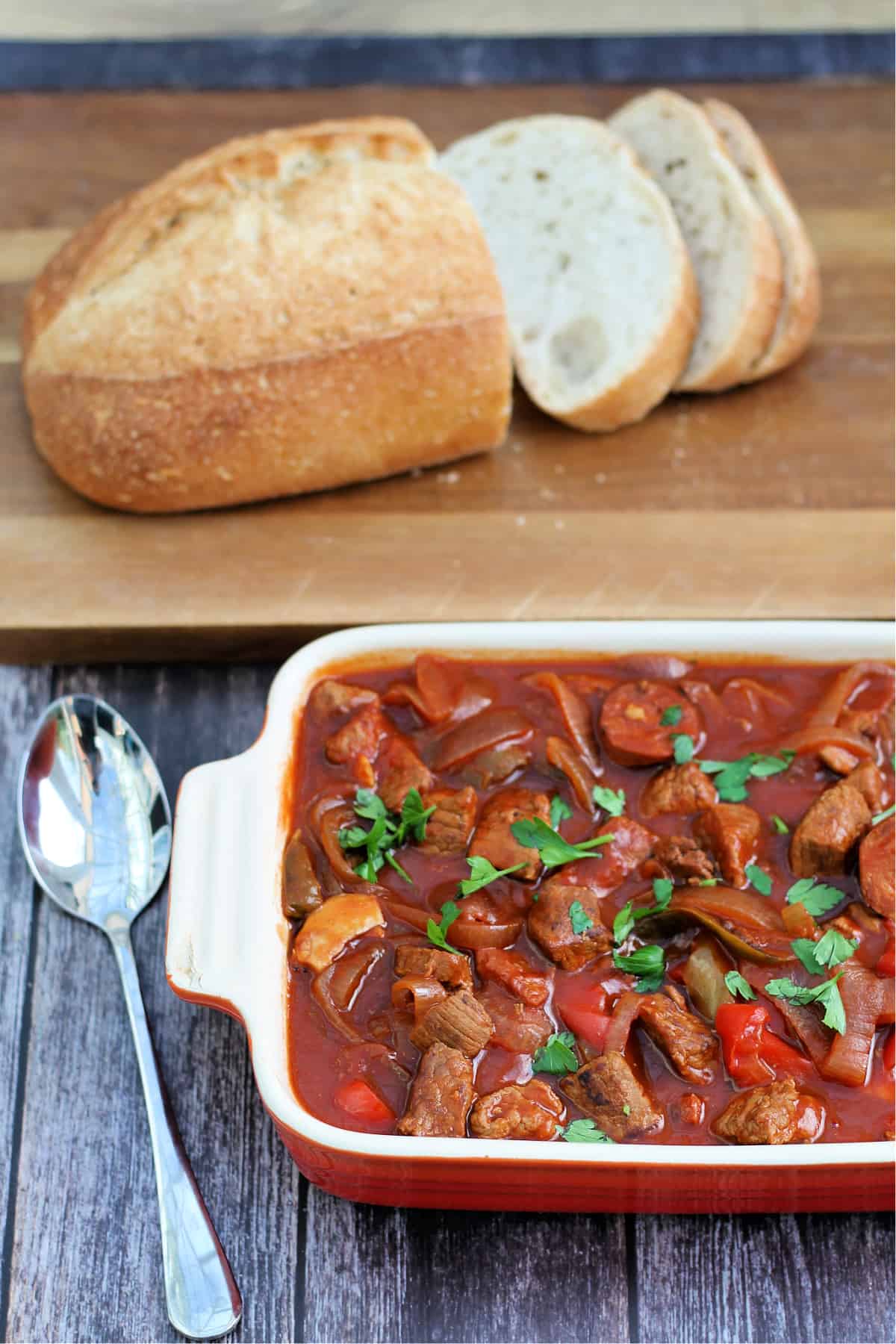 Serving dish of casserole with bread board with sliced crusty loaf behind.