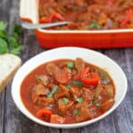 Bowl of beef casserole with bread on the side, with serving dish behind, with text overlay (slow cooker beef casserole).