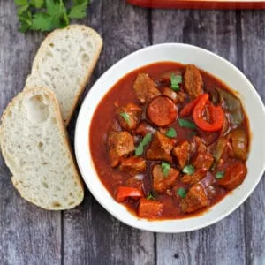 Bowl of beef casserole served with crusty bread on the side, with herbs and serving dish behind.