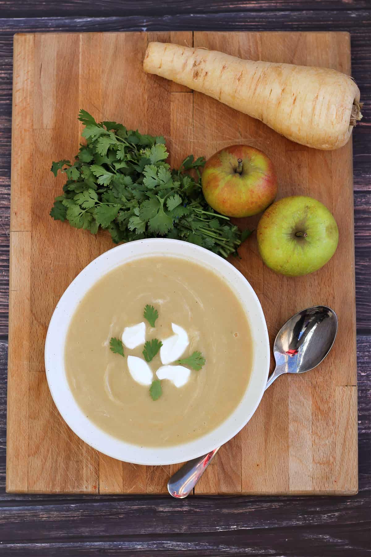 Bowl of soup on a wooden board with parsnip, apples and herbs behind.