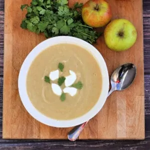 View from above of bowl of soup on wooden board with apples and herbs beside.