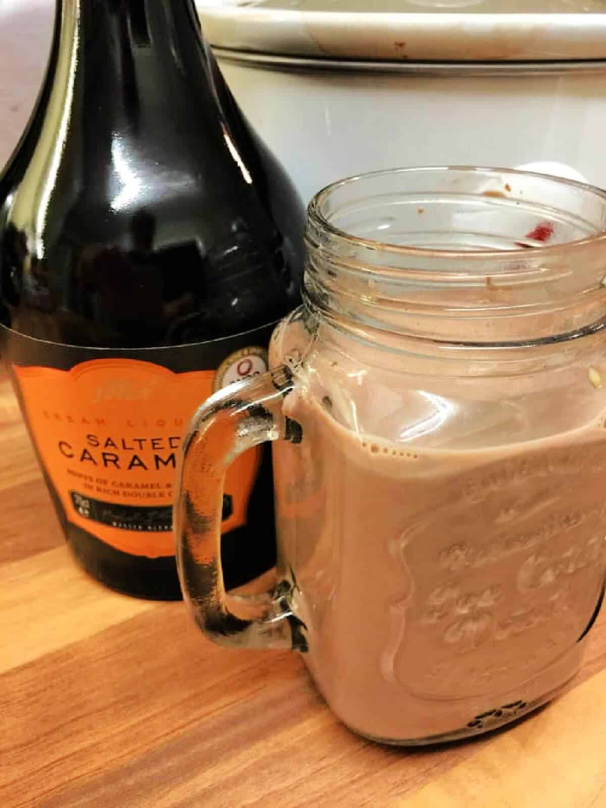 Glass mug of hot chocolate next to bottle of caramel liqueur.