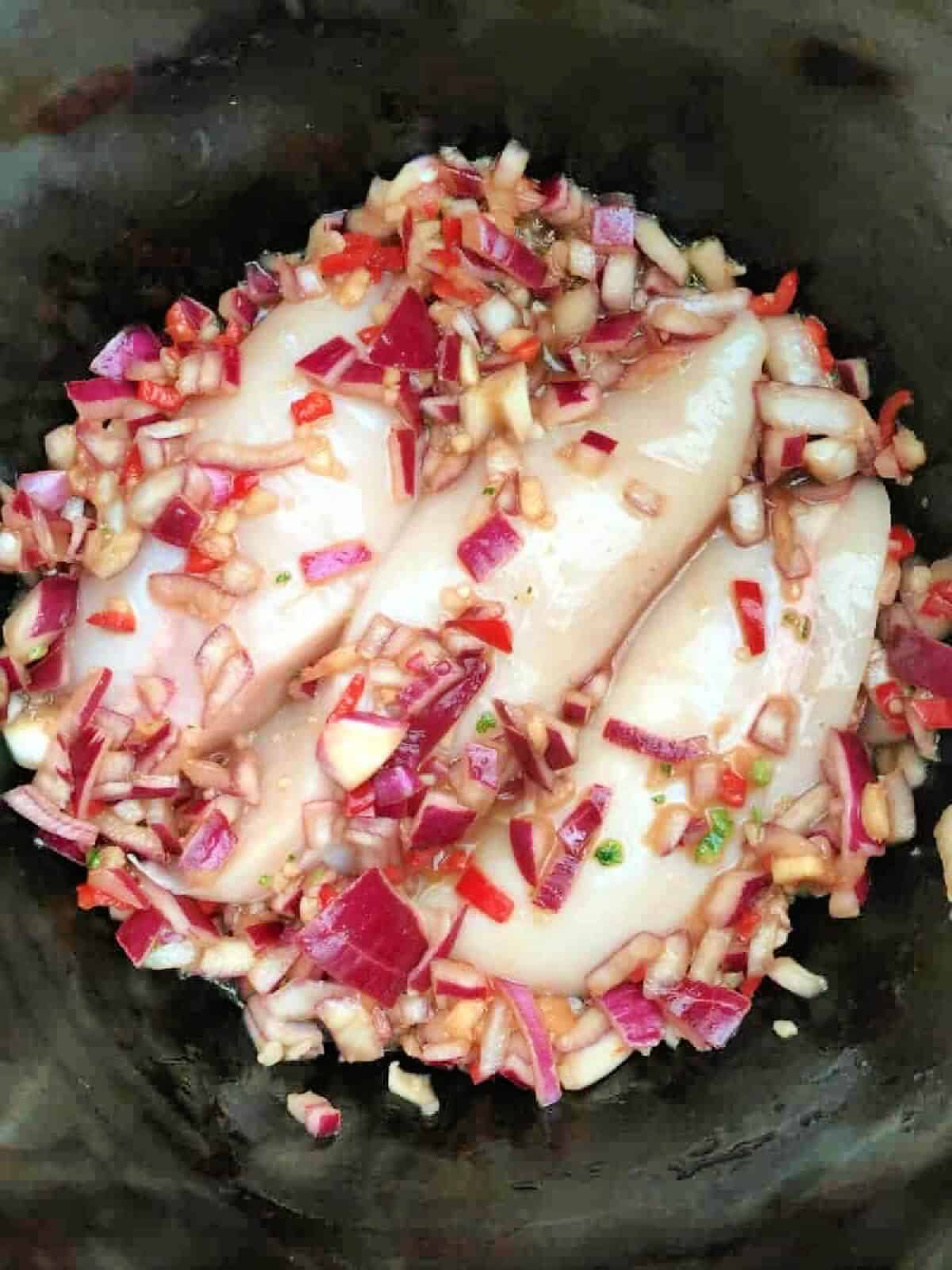 Chicken breasts with diced red onion and red chilli in slow cooker pot.