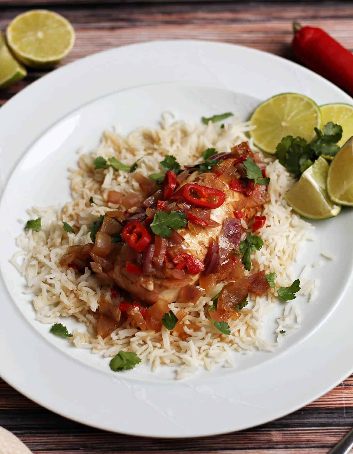 Chicken topped with red onion, red chilli and garnished with coriander on a white plate with rice and lime wedges.