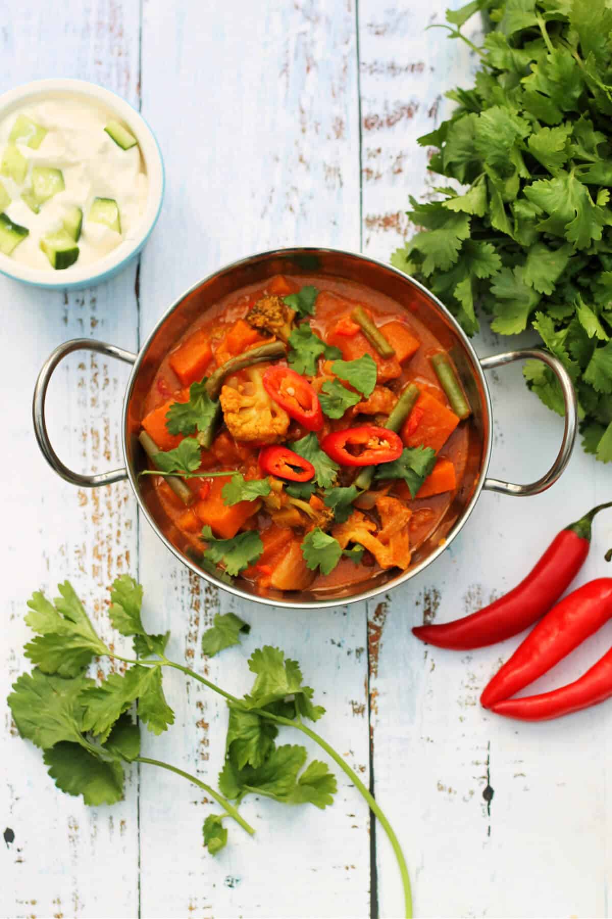 Overhead view of a bowl of cauliflower and sweet potato curry topped with red chilli and coriander, surrounded by ingredients.