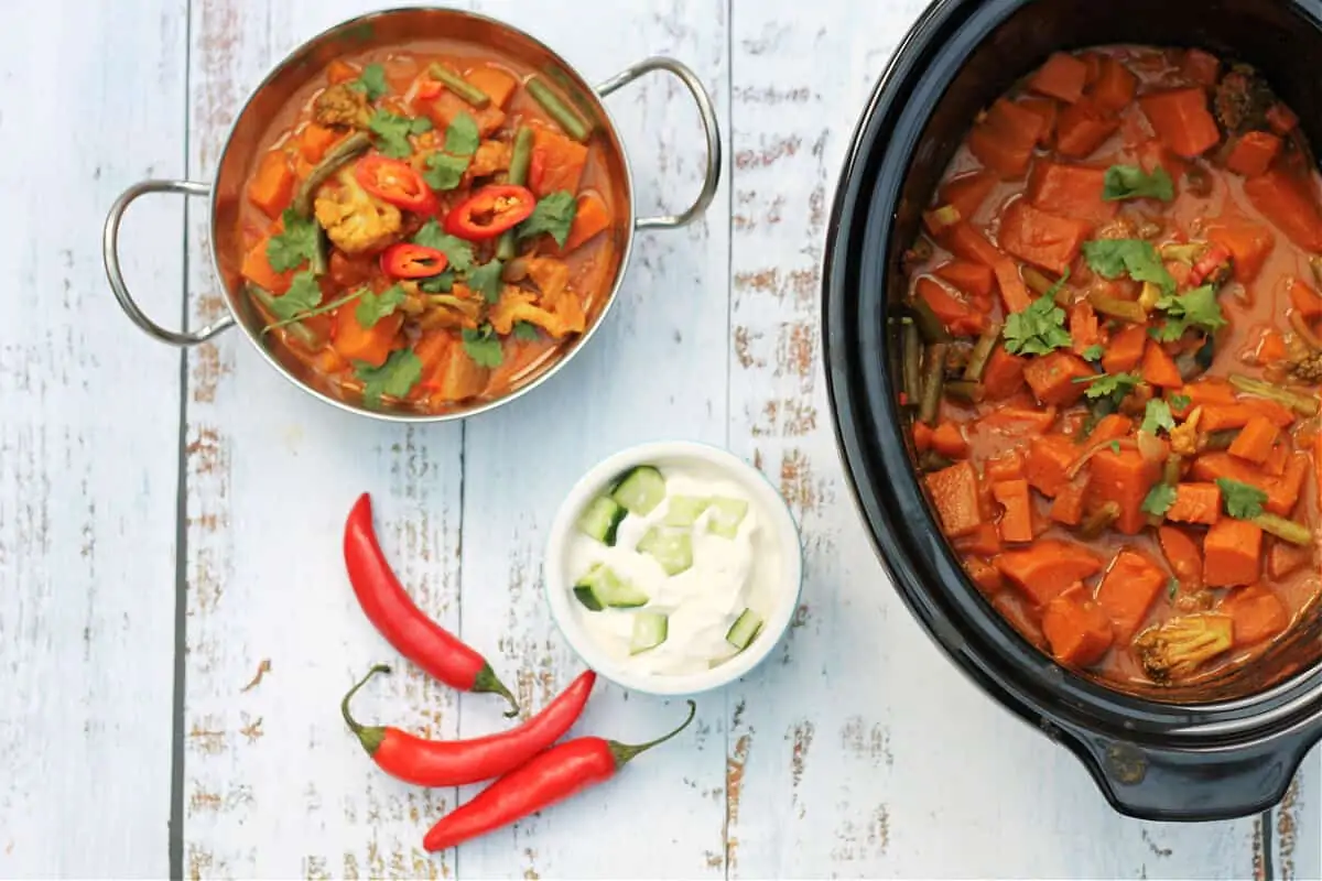 Serving dish of slow cooker pot with a bowl of curry to the side, plus a pot of raita and some red chillis.