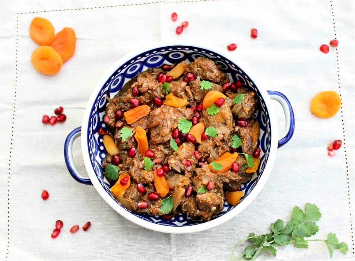 Lamb tagine in a blue and white serving bowl, with ingredients scattered on the tablecloth around it.