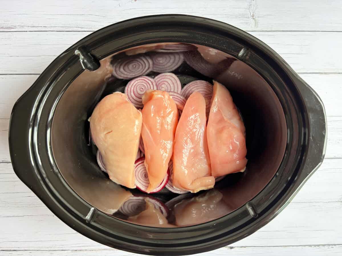 Chicken breasts and red onion slices in a slow cooker cooker pot.