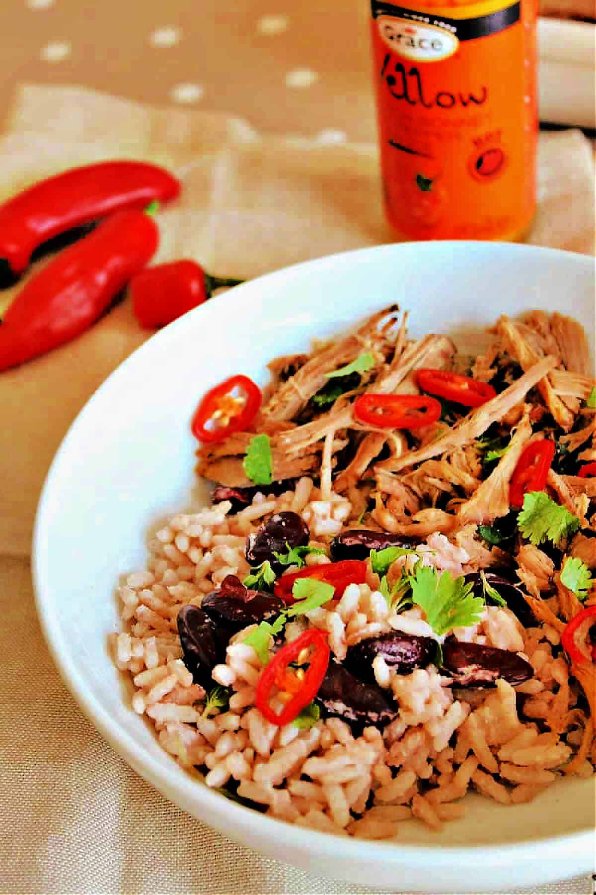 Pulled pork garnished with red chilli and coriander, served in a white bowl with rice and peas.
