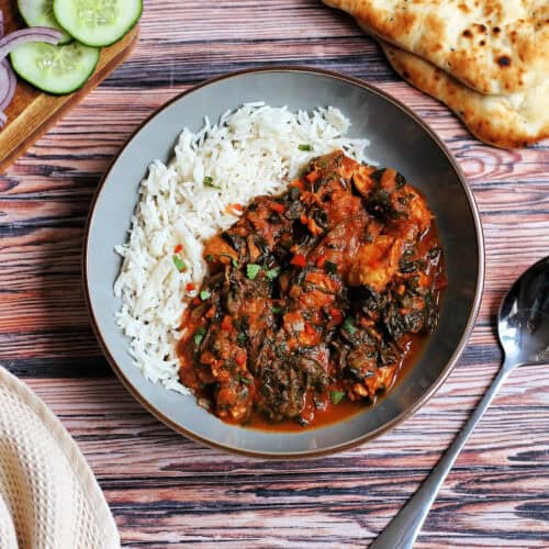 Chicken spinach curry in a grey bowl, spoon on one side, naan bread and cucumber on other sides.