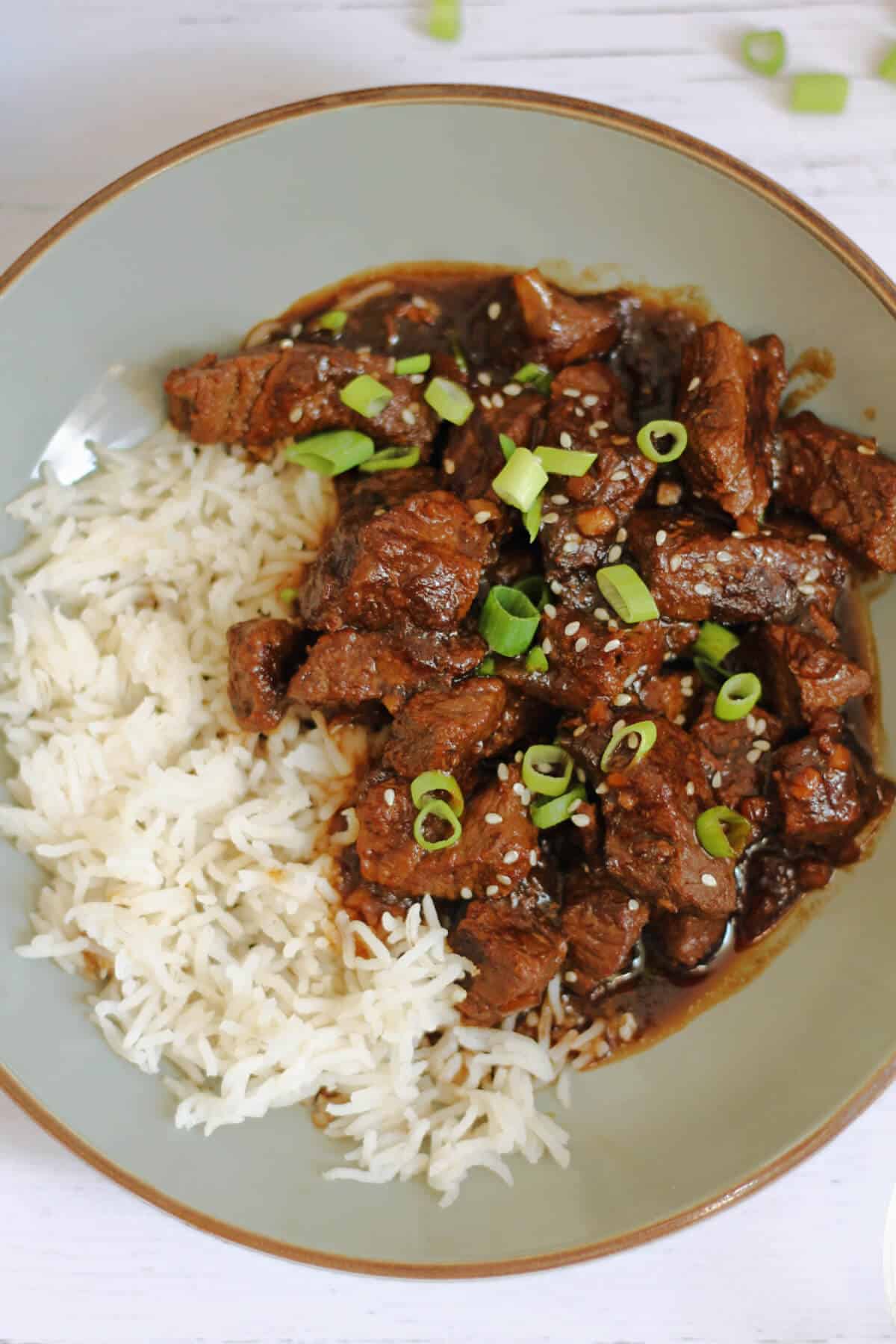 Teriyaki beef in a rich brown sauce garnished with sesame seeds and chopped spring onions, with rice in a grey bowl.