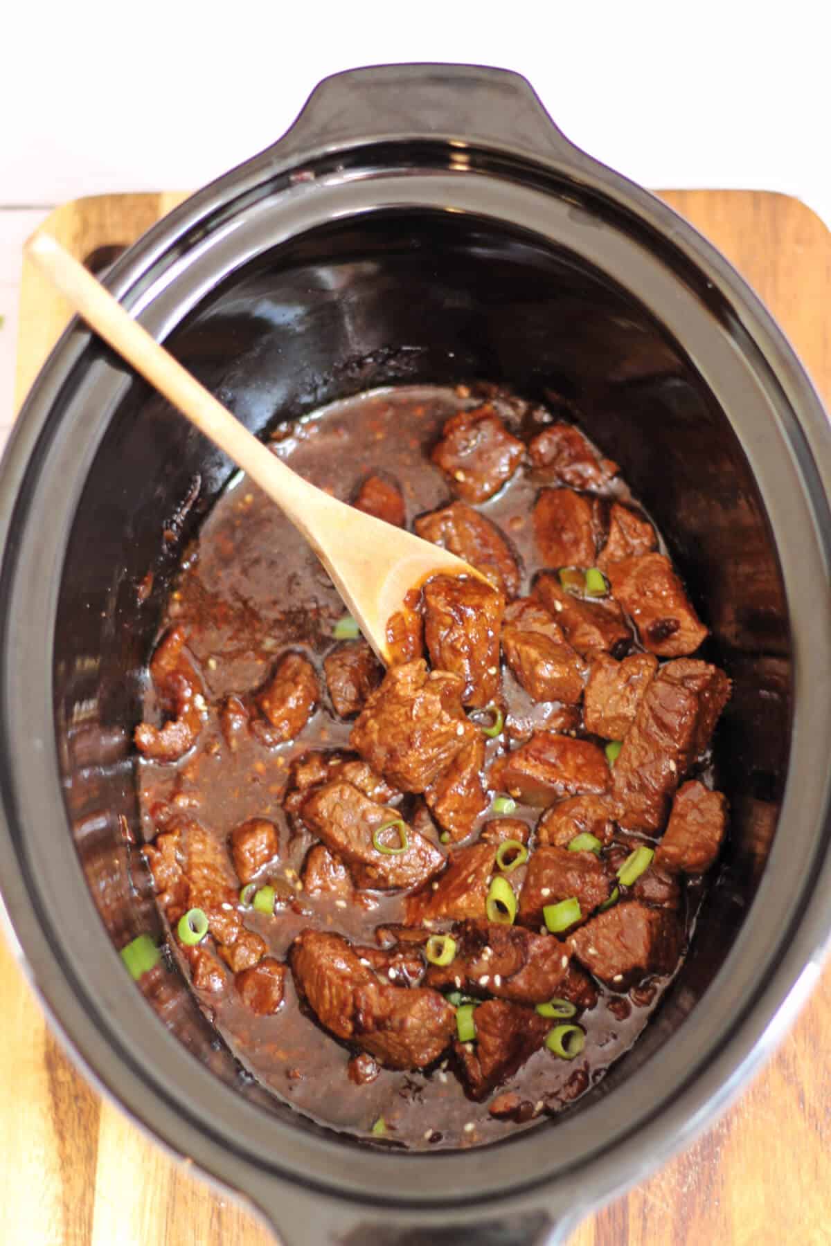 Slow cooker pot with beef in a rich glossy sauce, with wooden spoon in it.