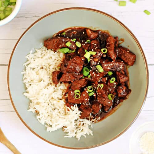 Teriyaki beef in a rich brown sauce garnished with sesame seeds and chopped spring onions, with rice in a grey bowl.