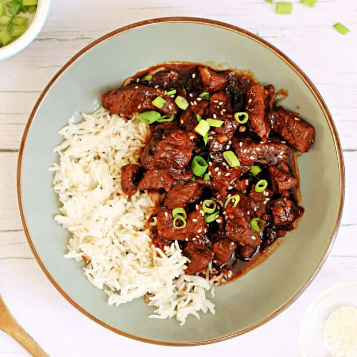 Teriyaki beef in a rich brown sauce garnished with sesame seeds and chopped spring onions, with rice in a grey bowl.