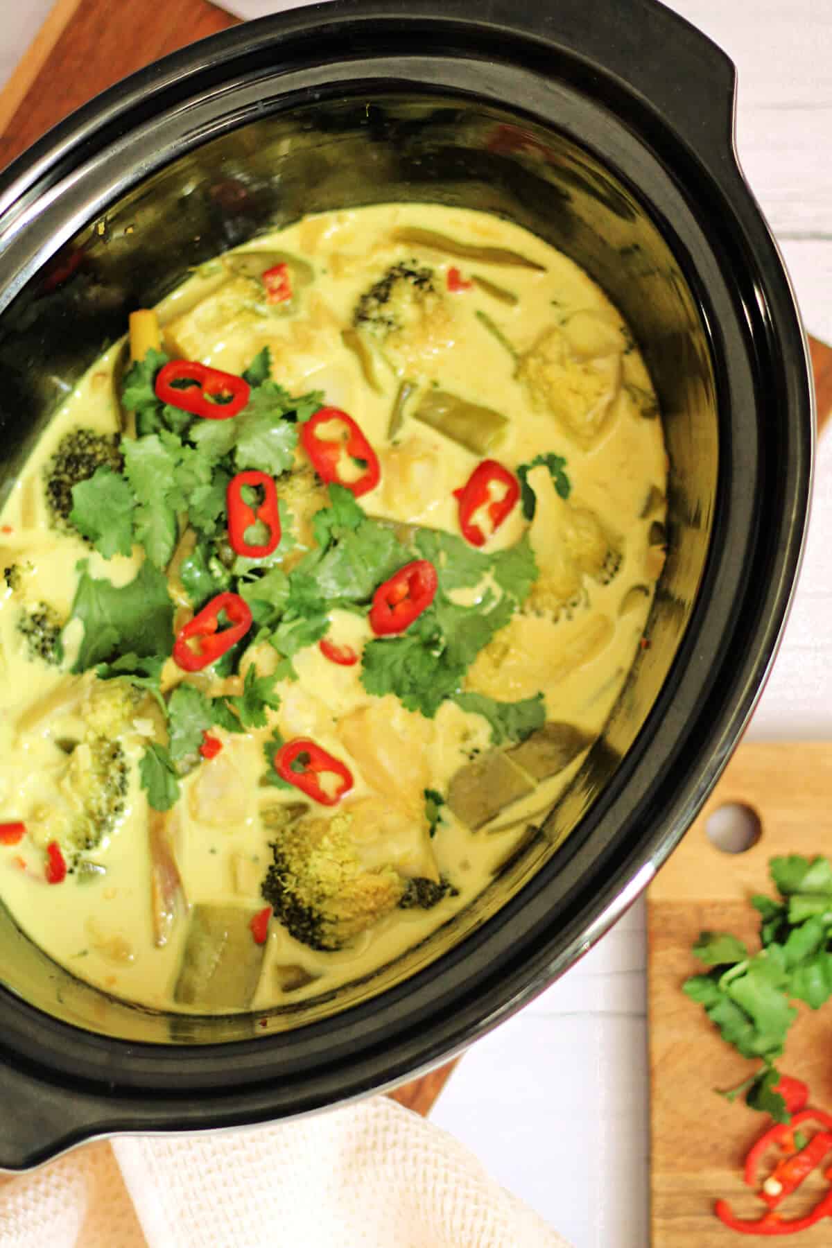 Top view of slow cooker pot with yellow fish curry inside, chopped herbs to the side on a board.
