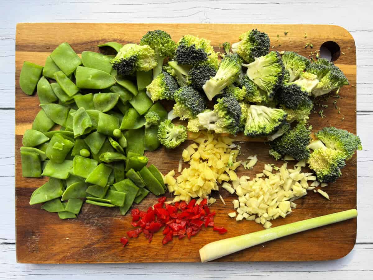 Chopped mange tout, brocoli, garlic, ginger, red chilli and a stick of lemongrass on a board.