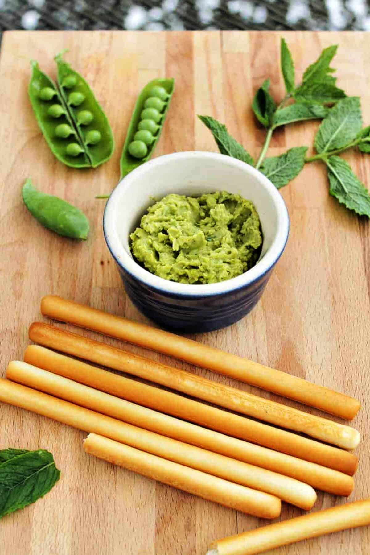 Pot of broad bean hummus with broad bean pods and mint leaves on a board.