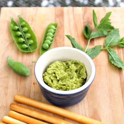 Pot of green hummus on a wooden board, pea pods behind.