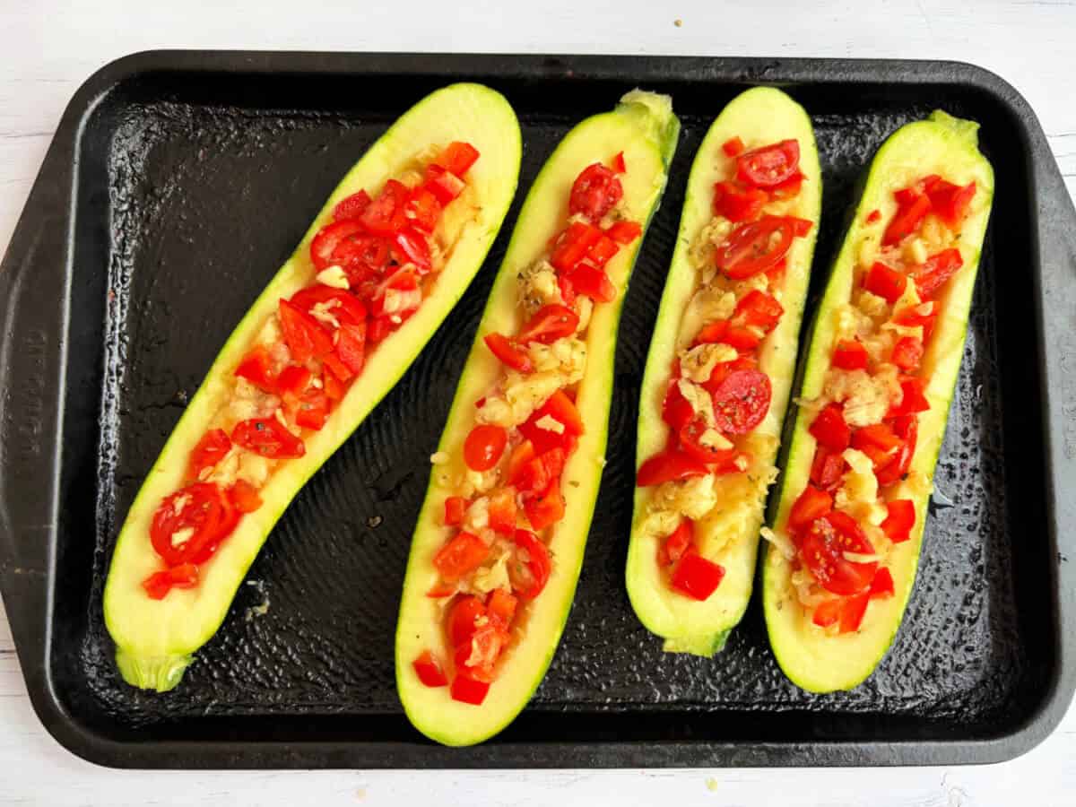 Courgette halves on baking tray with pepper and tomato filling added.