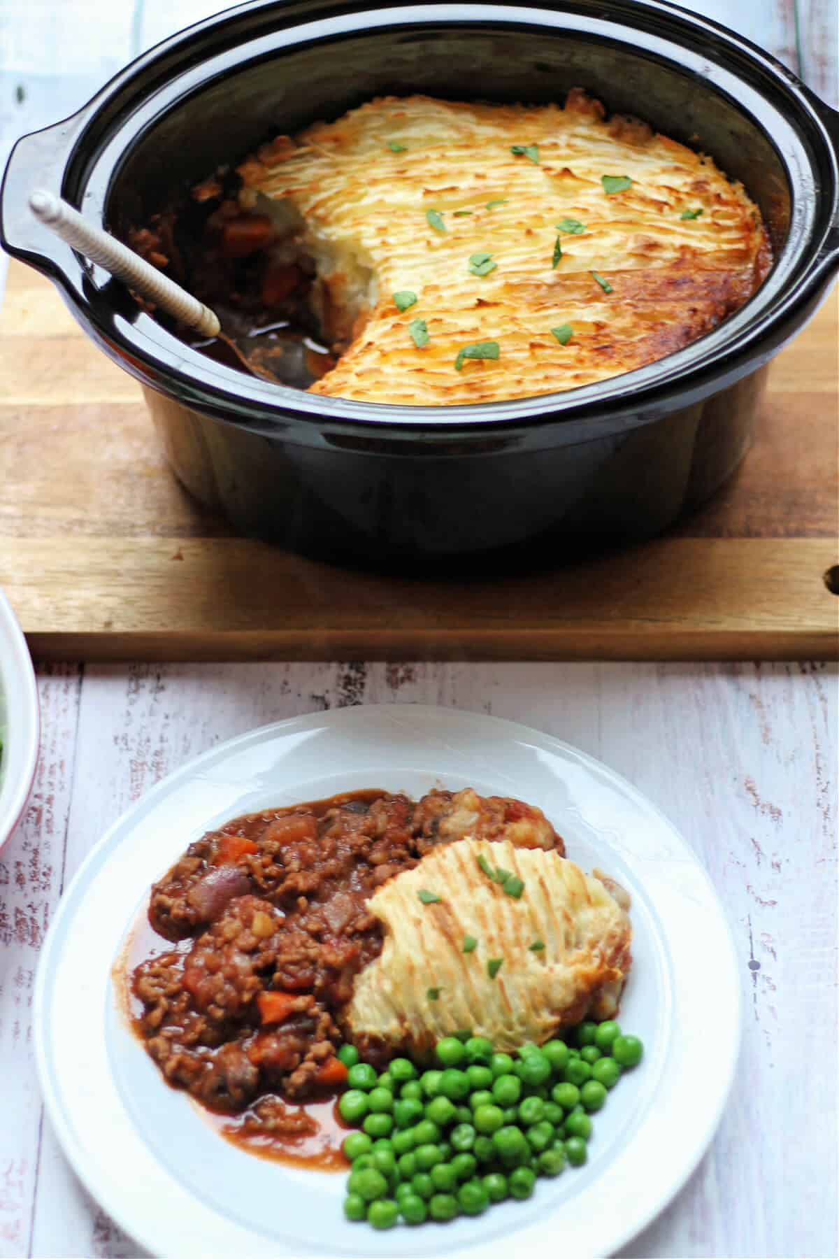 Plate of cottage pie served with peas, with serving dish behind.