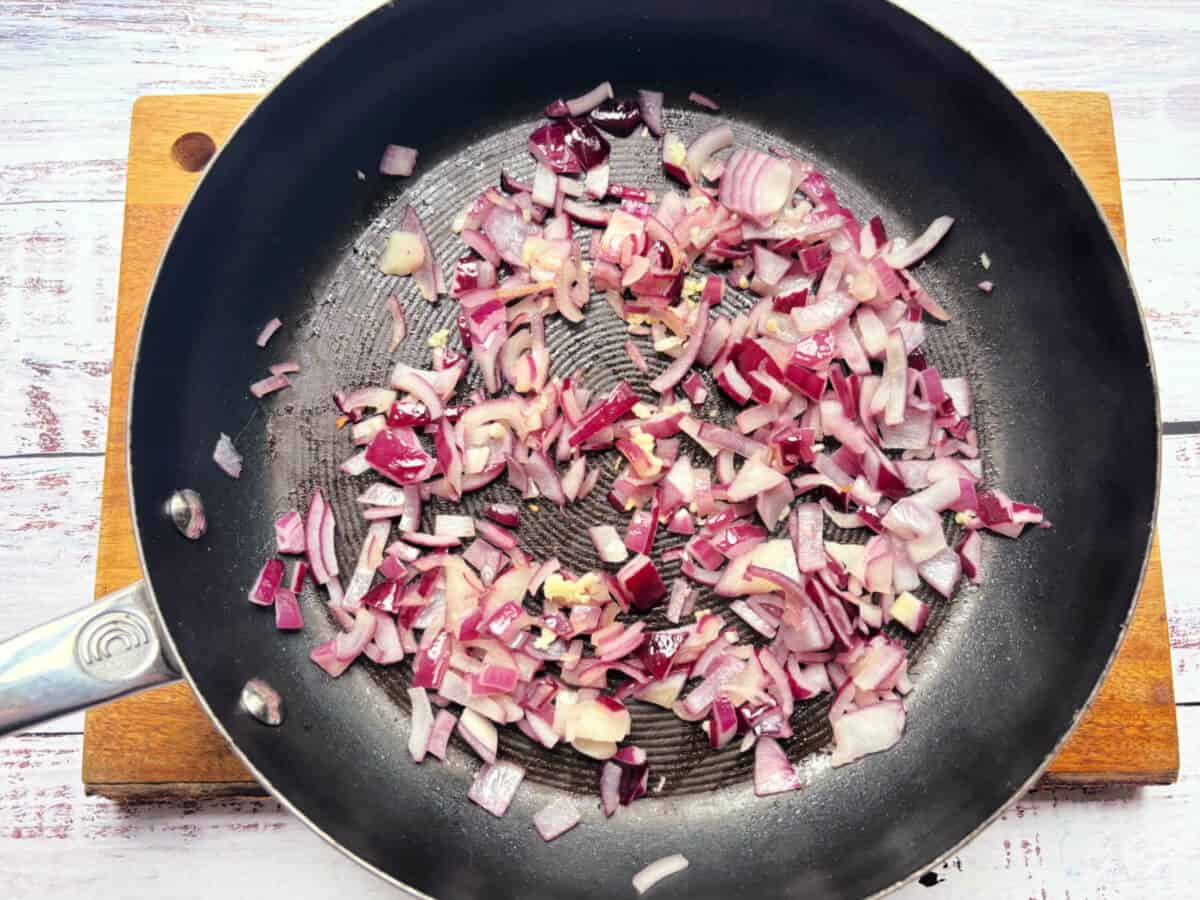 Chopped red onion and garlic being fried in a frying pan.
