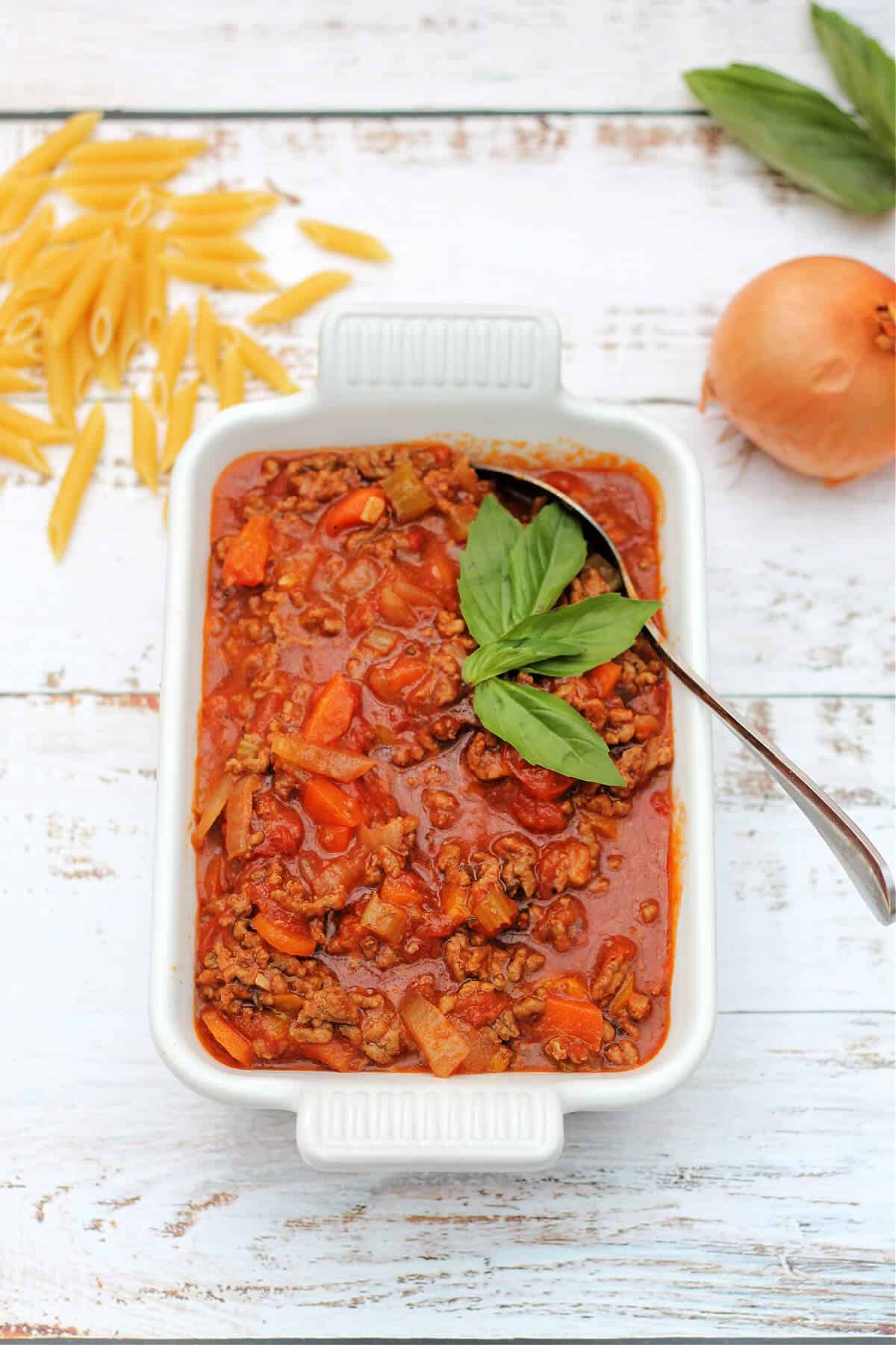Bolognese sauce served in a white serving dish, with pasta and onion in the background.